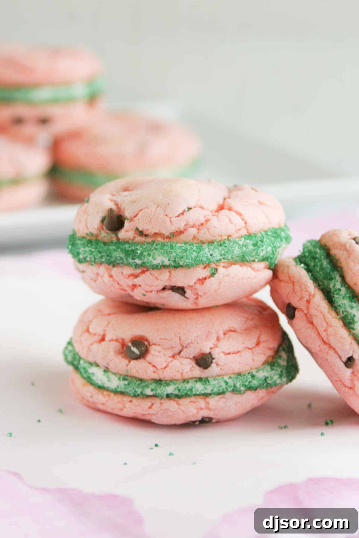 These Watermelon Cake Mix Cookie Sandwiches look like summer! Strawberry cake mix cookies are studded with chocolate chips, filled with buttercream frosting, and rolled in green sugar. Stacked Watermelon Cake Mix Cookie Sandwiches with vibrant green edges and chocolate chip 'seeds'