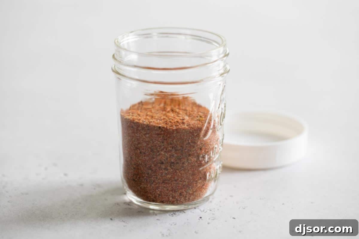 A small glass jar filled with golden-brown homemade Cajun seasoning, sitting on a wooden surface.