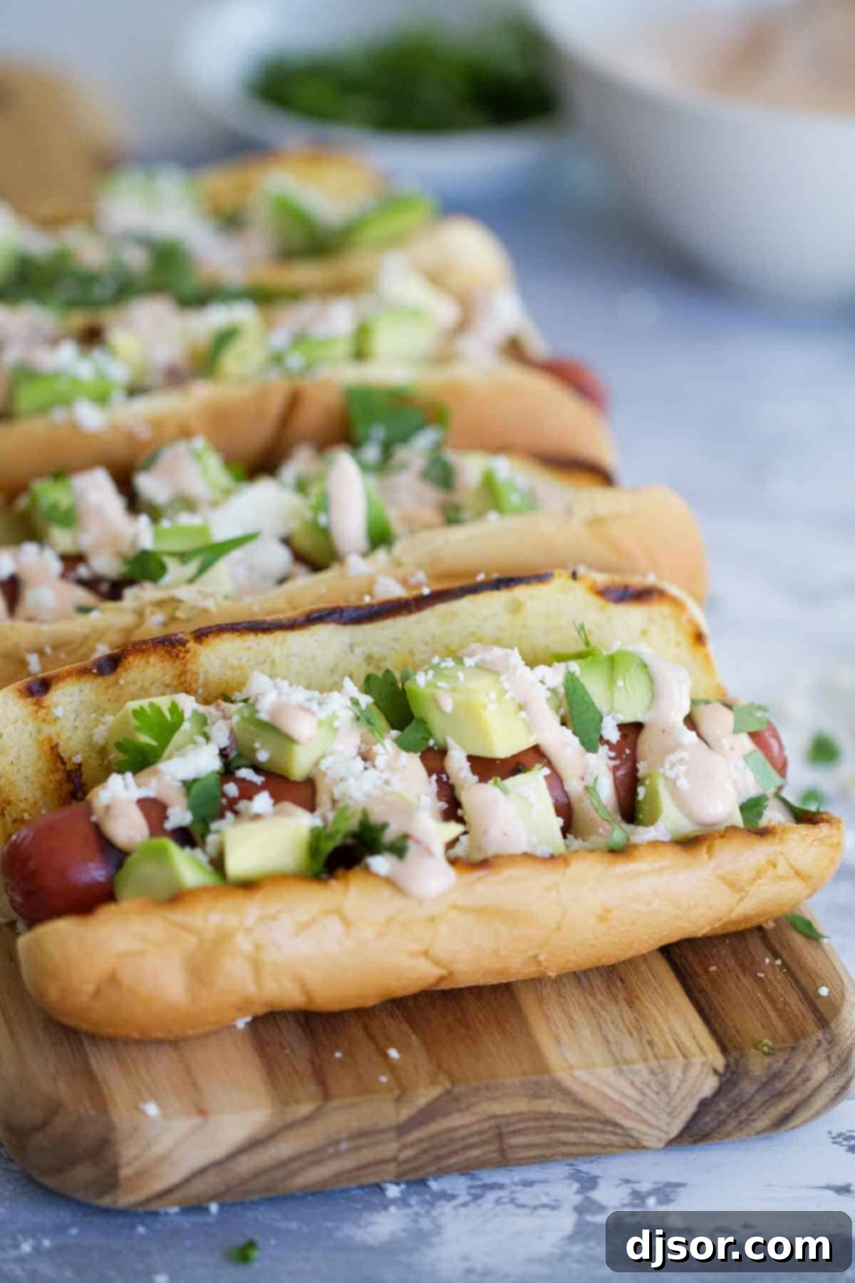 Hot dogs with Tex-Mex flair - this Mexican Hot Dog recipe gives you hot dogs topped with a chipotle mayonnaise, avocados, cheese and cilantro. Close-up view of freshly prepared Mexican Hot Dogs arranged on a wooden cutting board, garnished with vibrant toppings.