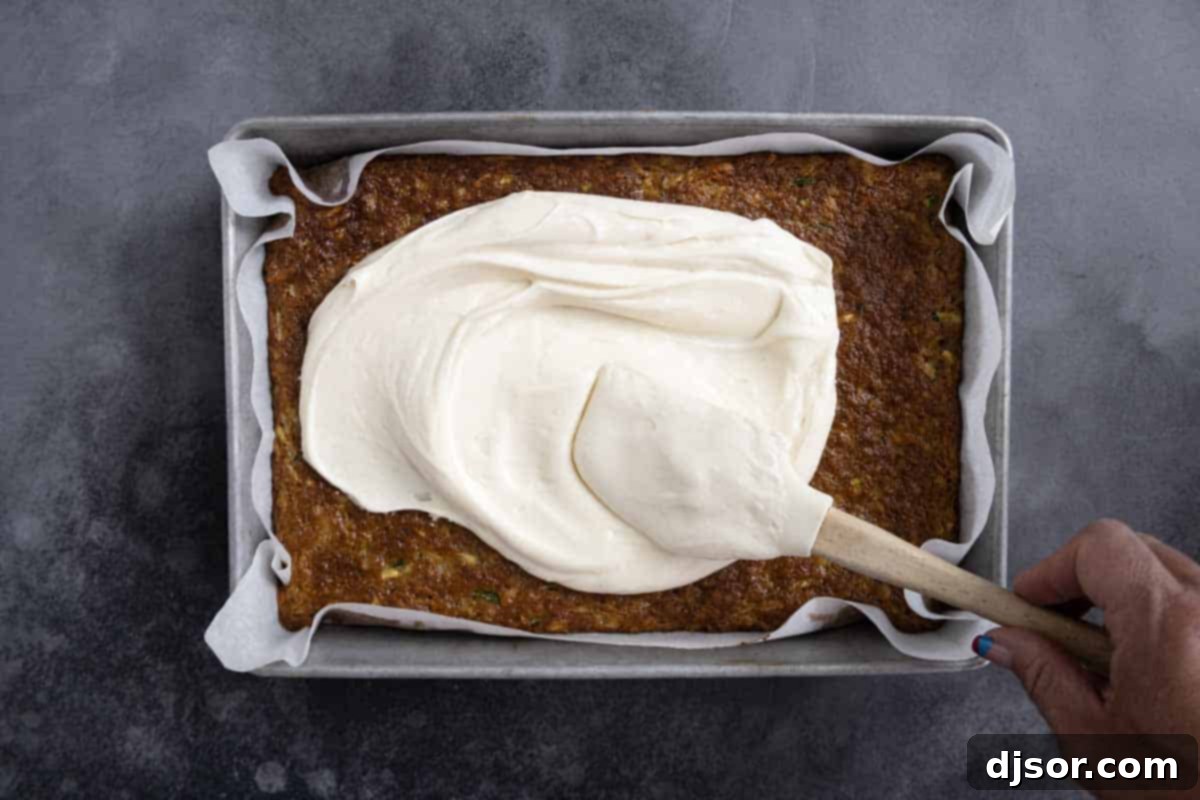A baker's hand delicately spreading smooth cream cheese frosting over the cooled Zucchini Cake in a baking pan.