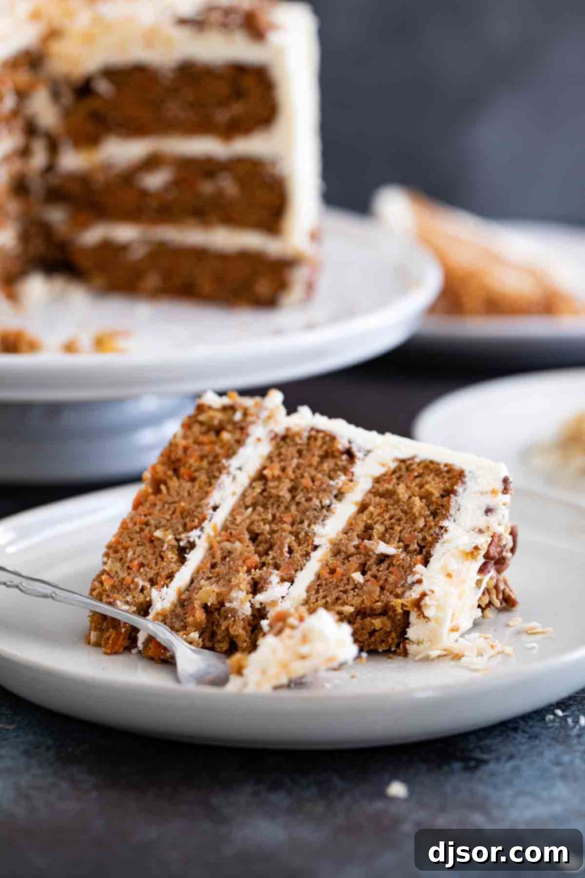 A beautiful slice of perfectly moist traditional carrot cake topped with creamy white cream cheese frosting, with a full three-layer carrot cake in the soft-focus background, ready to be served.