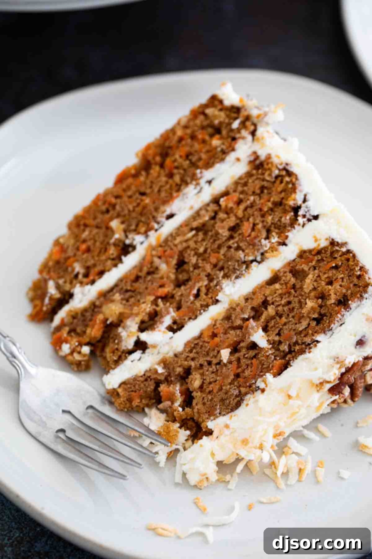 A close-up view of a slice of traditional layered carrot cake on a white plate, showcasing its moist texture, visible carrot bits, and thick cream cheese frosting layers.