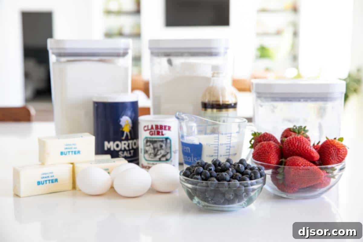 Ingredients laid out on a table to make a flag cake, including flour, sugar, butter, eggs, milk, vanilla, baking powder, salt, blueberries, and strawberries.