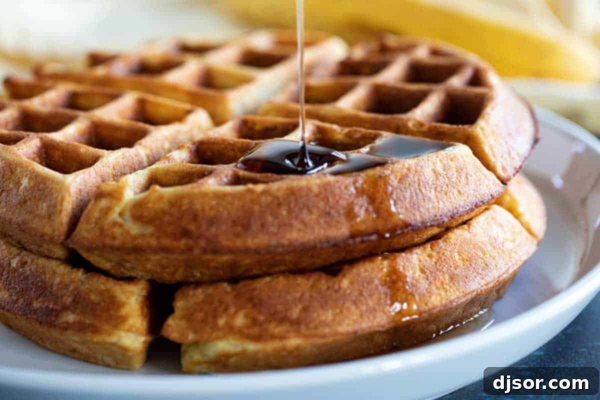Stack of Banana Waffles with Maple Syrup A beautiful stack of two golden-brown banana waffles sits on a white plate, with a generous stream of maple syrup being poured over the top, creating a tempting breakfast scene.