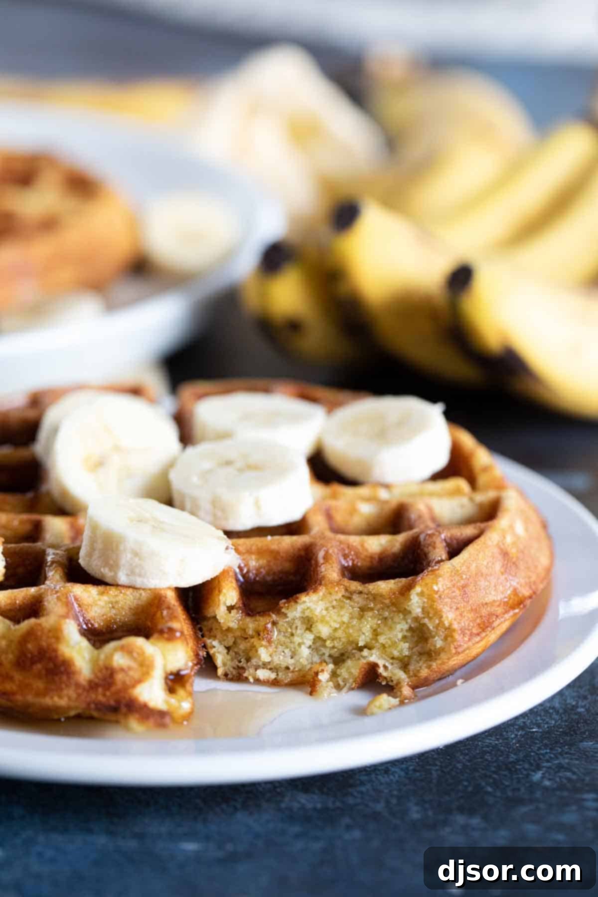 Close-Up of Banana Waffle Texture A close-up shot of a banana waffle on a plate, with a bite taken out to reveal its fluffy, airy interior and the visible pieces of mashed banana within. It's topped with fresh banana slices and a light dusting of powdered sugar.
