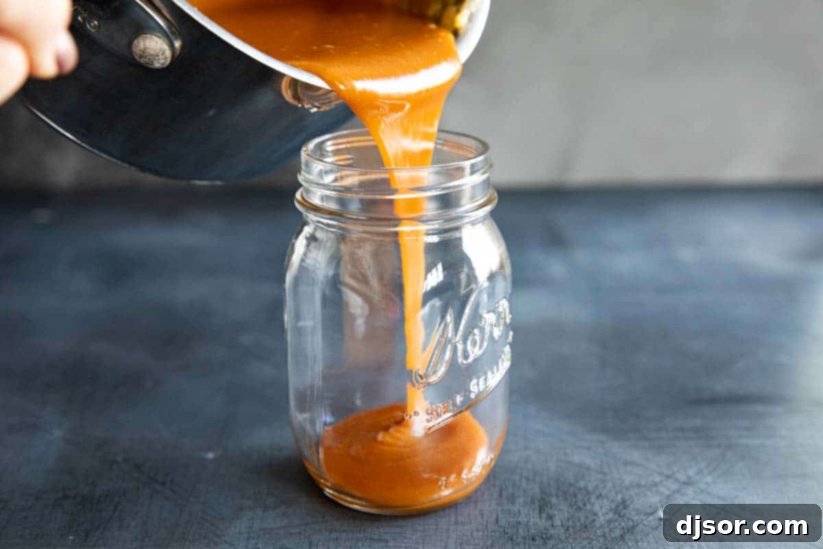Hot, freshly made salted caramel sauce being poured from a saucepan into a clear glass storage jar.