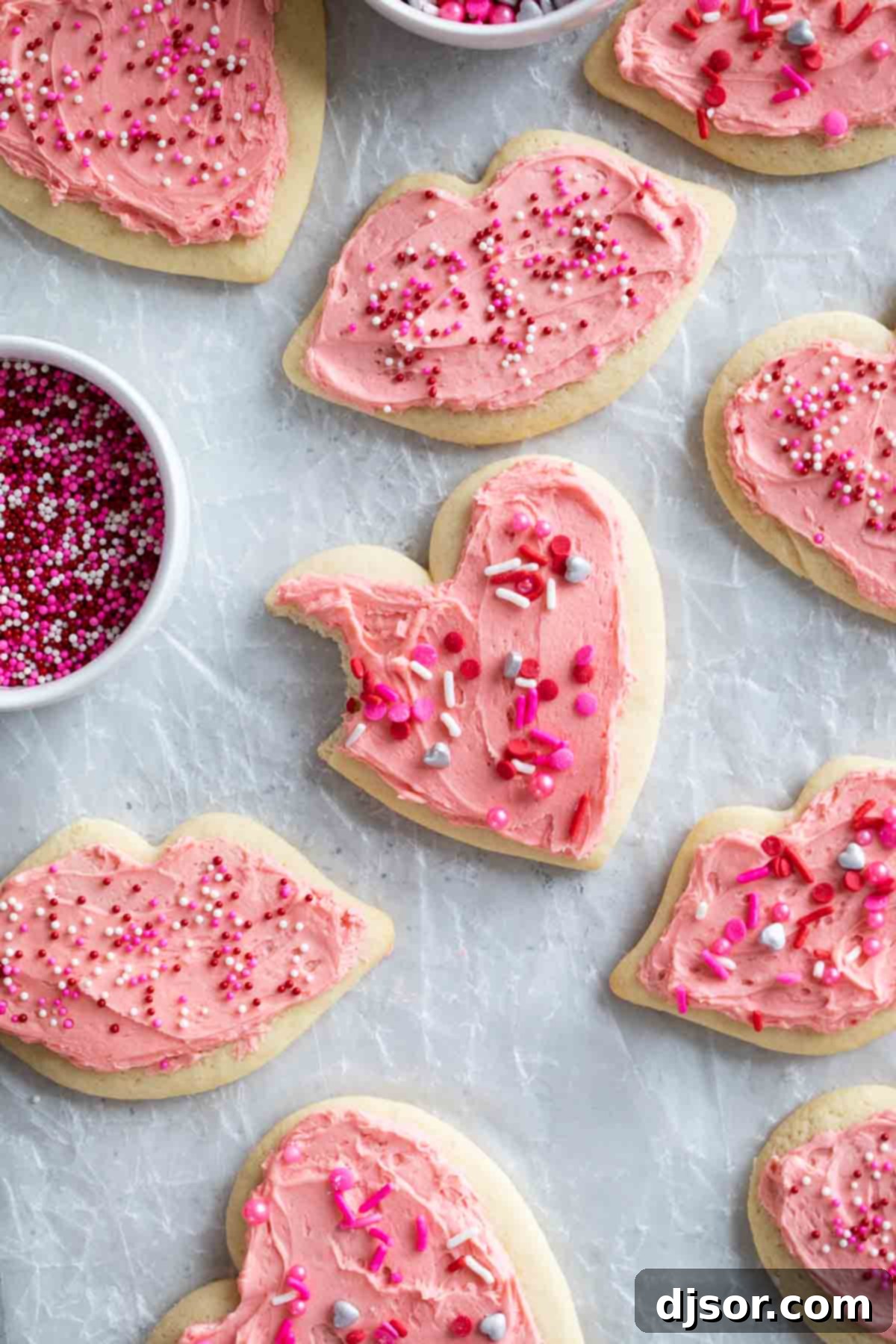 Pretty in pink, these Valentine's Day Sugar Cookies start with an easy sugar cookie recipe that is topped with the perfect pink buttercream and lots of holiday sprinkles. Valentine's Day Sugar Cookies on parchment with a bite taken from one cookie.
