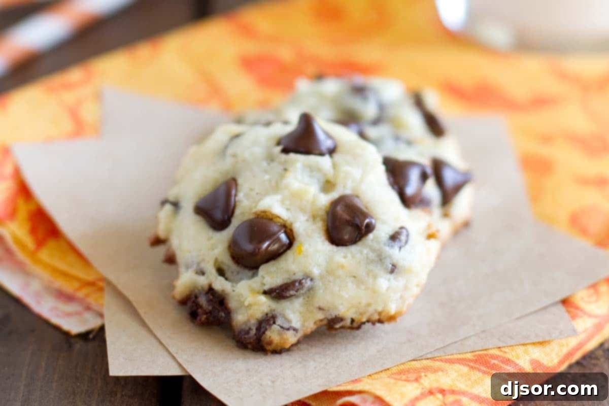 Two freshly baked Orange Chocolate Chip Cookies on parchment paper, showcasing their soft centers and golden edges, with a scattering of orange zest in the background.