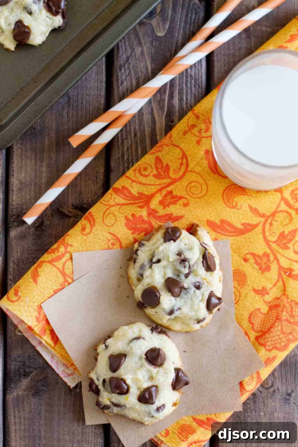 A close-up of two Orange Chocolate Chip Cookies on an orange fabric, highlighting their perfect golden-brown edges and abundance of melted chocolate chips.