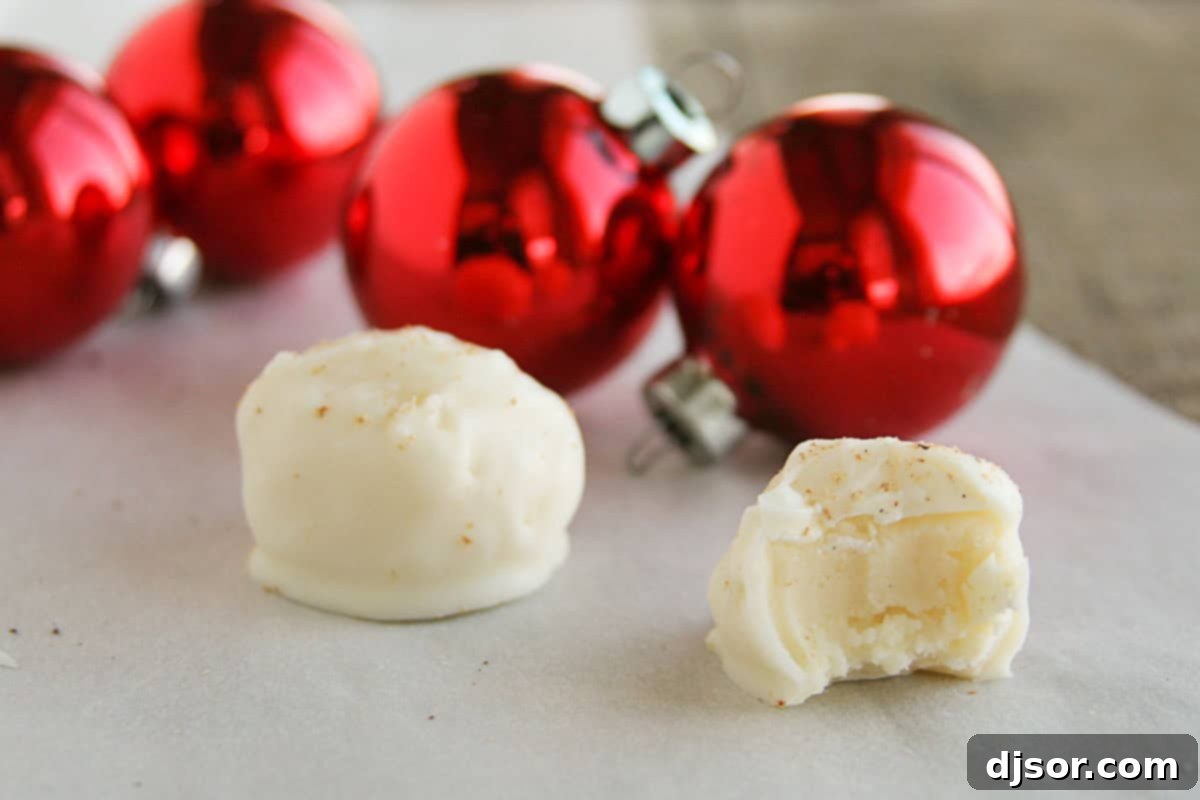 Two luxurious eggnog truffles, one with a delicate bite taken, revealing its creamy white chocolate interior. Sprinkled with nutmeg.