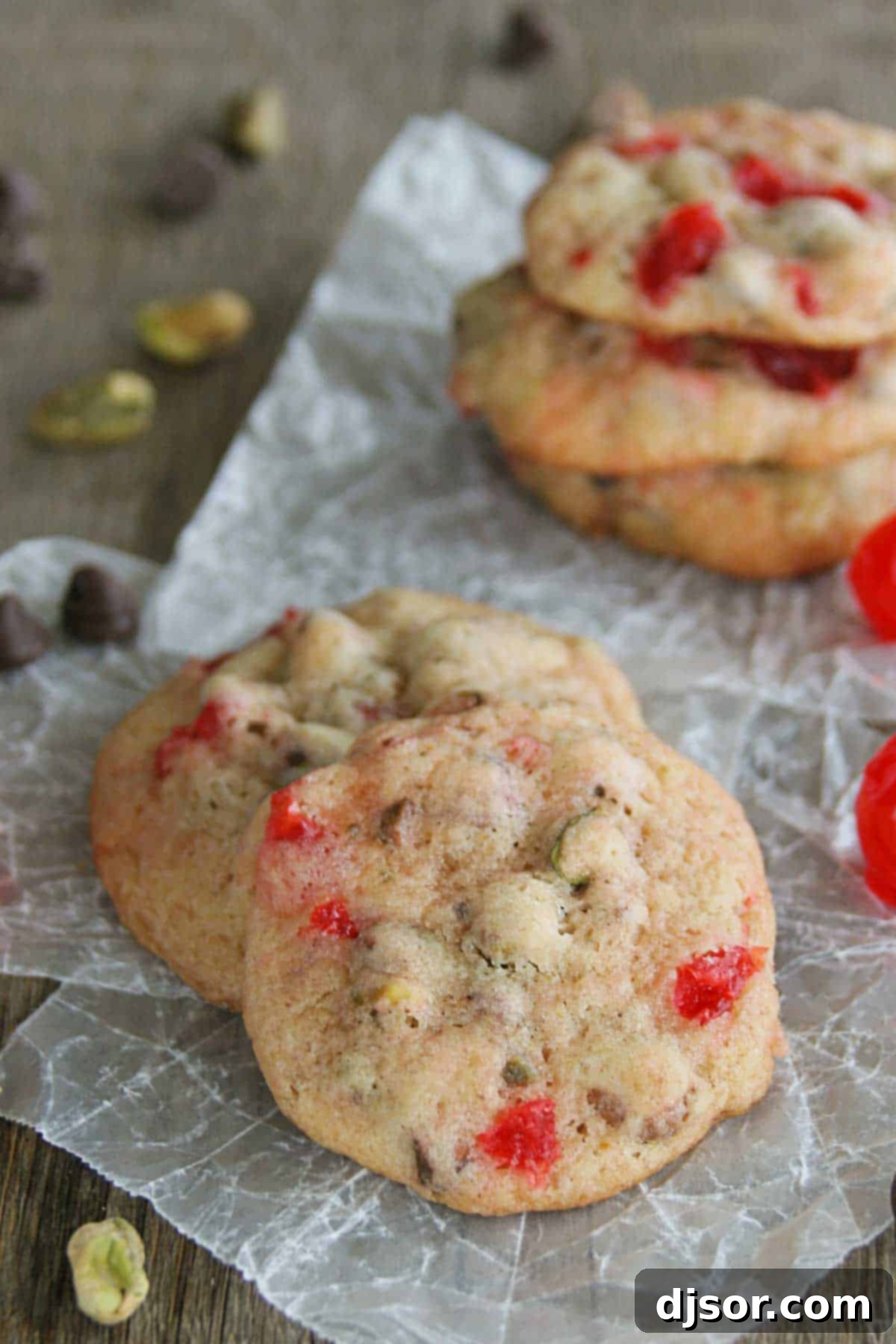 Cherry Chocolate Nut Cookies sitting on each other, with pistachios, cherries, and chocolate chips surrounding them, creating a festive display.