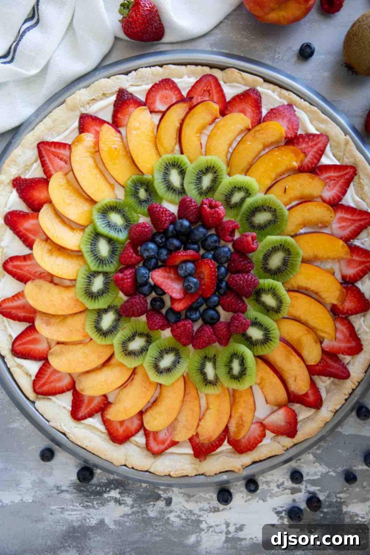 Indulge in the sweetness of summer with this irresistible Fruit Pizza. A sugar cookie base is topped with a sweet cream cheese frosting, then layered with fresh, in season fruit for a stunning dessert that is as delicious as it beautiful. Fruit pizza consisting of a sugar cookie base topped with cream cheese frosting and fresh fruit.