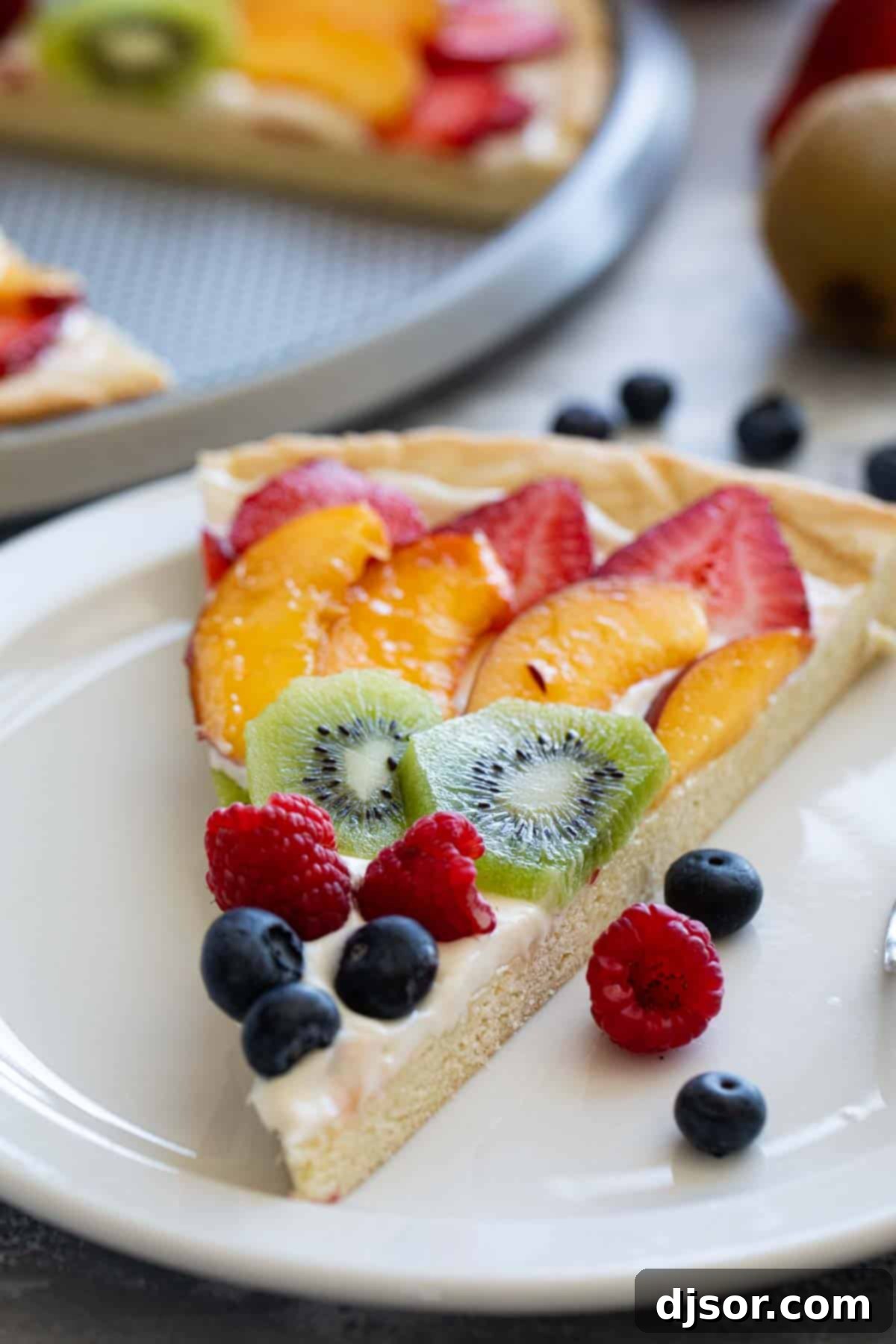 Indulge in the sweetness of summer with this irresistible Fruit Pizza. A sugar cookie base is topped with a sweet cream cheese frosting, then layered with fresh, in season fruit for a stunning dessert that is as delicious as it beautiful. Slice of fruit pizza on a plate.