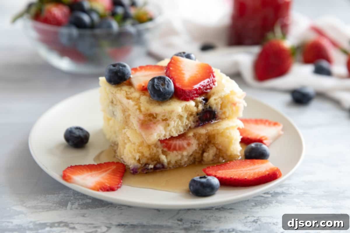 Two slices of sheet pan pancakes on top of each other, with syrup, strawberry slices, and blueberries.
