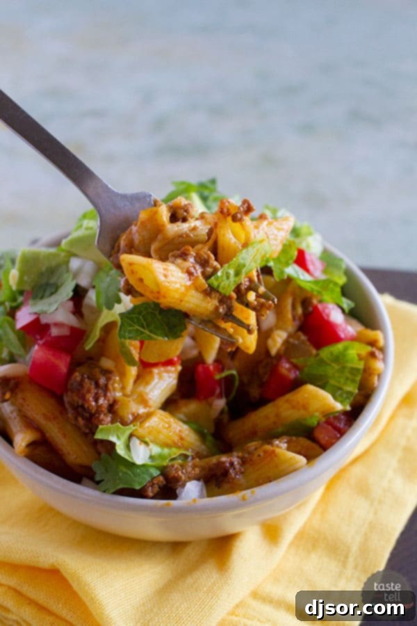 A close-up of a bowl of freshly prepared Taco Pasta Toss, garnished with a generous sprinkle of shredded cheddar cheese, diced fresh tomatoes, and vibrant green cilantro. The melty cheese over the pasta highlights the dish's comforting appeal.