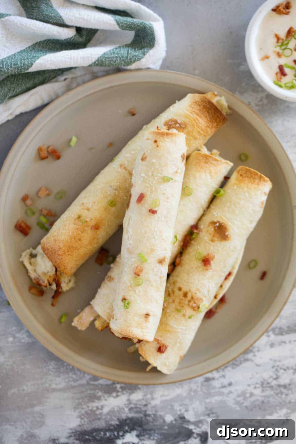 Close-up of freshly Baked Chicken Bacon Ranch Taquitos on a white plate, garnished with chopped green onions and a sprinkle of coarse salt.
