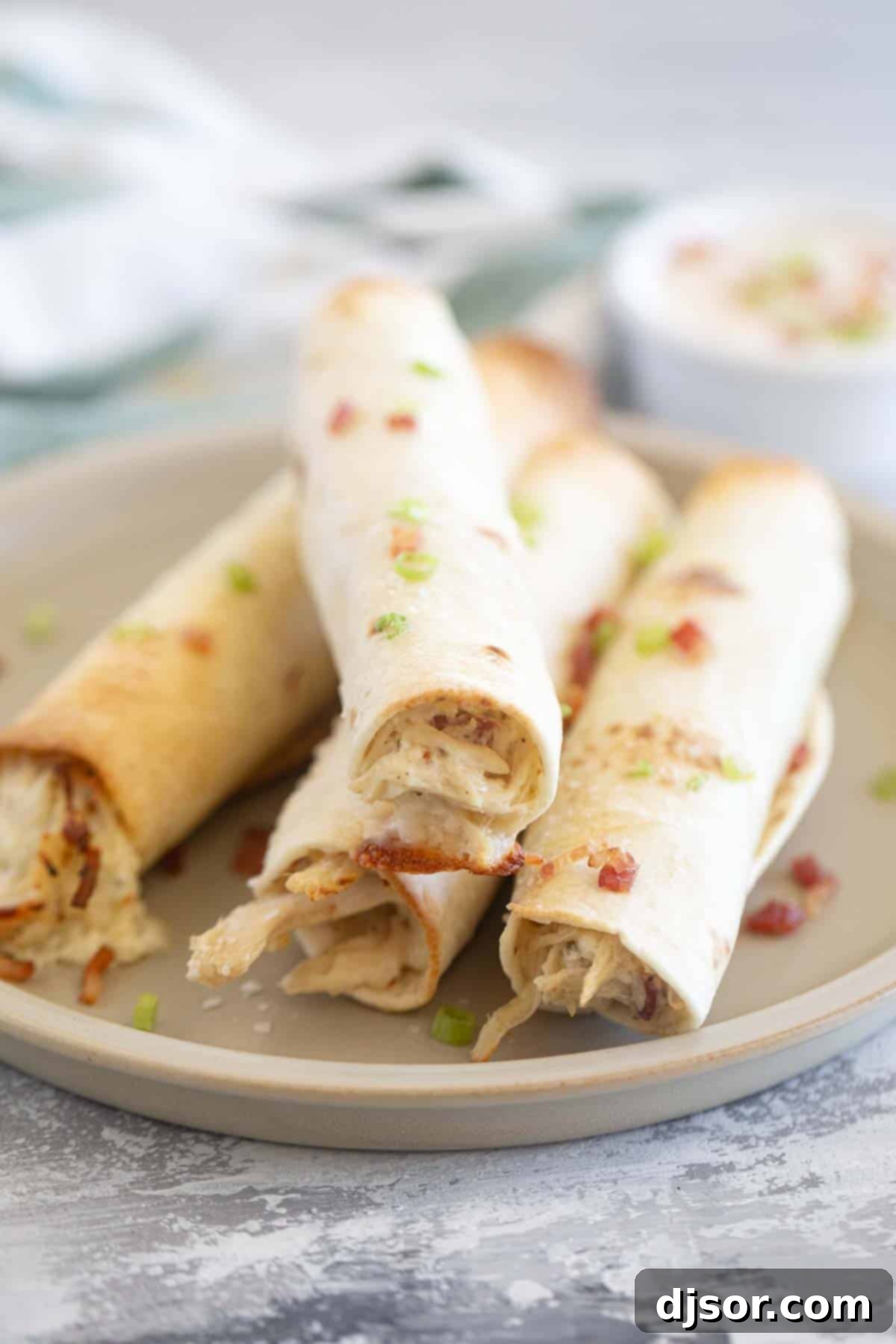 A stack of crispy Baked Chicken Bacon Ranch Taquitos on a white plate, garnished with fresh green onions.