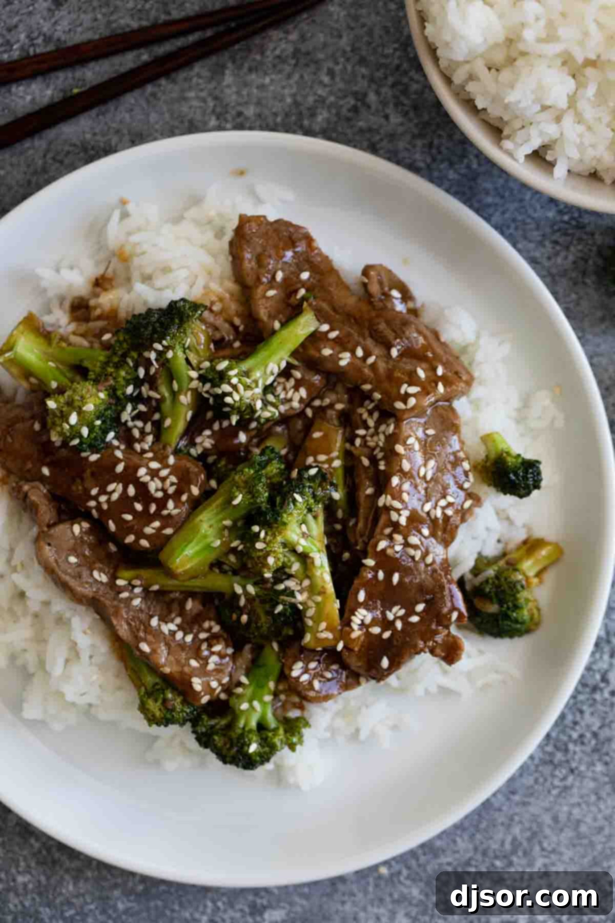 A serving of beef and broccoli over rice, garnished with toasted sesame seeds on a white plate.