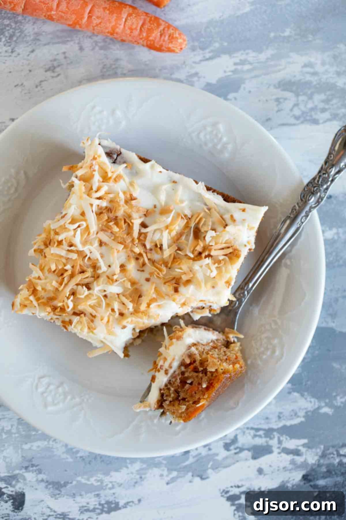 An inviting overhead view of a single slice of carrot sheet cake, generously frosted and topped with toasted coconut, presented on a white plate with a fork.