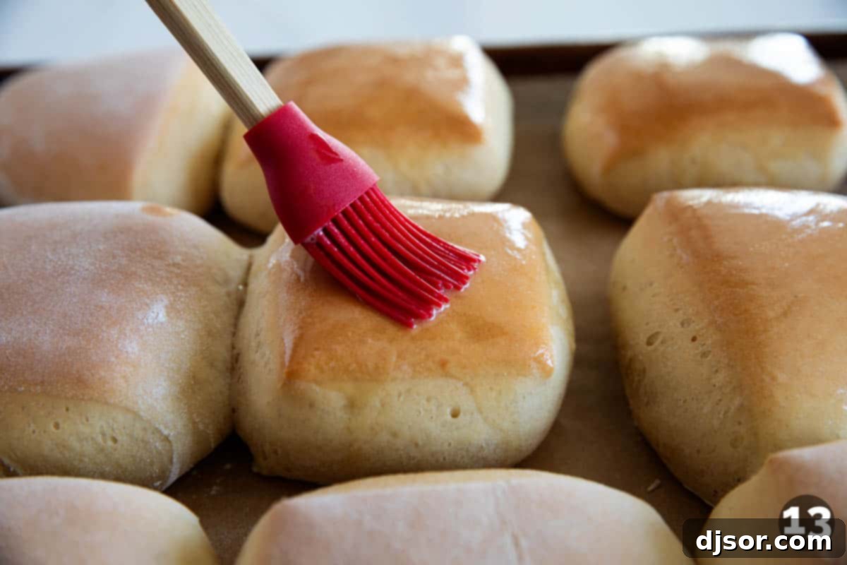 A close-up shot of a hand gracefully brushing melted butter over the tops of freshly baked, steaming Texas Roadhouse Rolls, creating a glossy and flavorful finish.