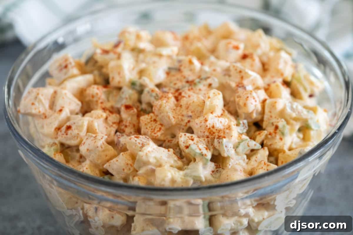 Classic Creamy Potato Salad 2 A large, inviting glass bowl filled to the brim with creamy, homemade potato salad, garnished with a sprinkle of paprika for a vibrant touch. The tender potato chunks, visible hard-boiled egg pieces, and fresh green celery bits are perfectly coated in the rich dressing, making it an irresistible centerpiece for any meal.