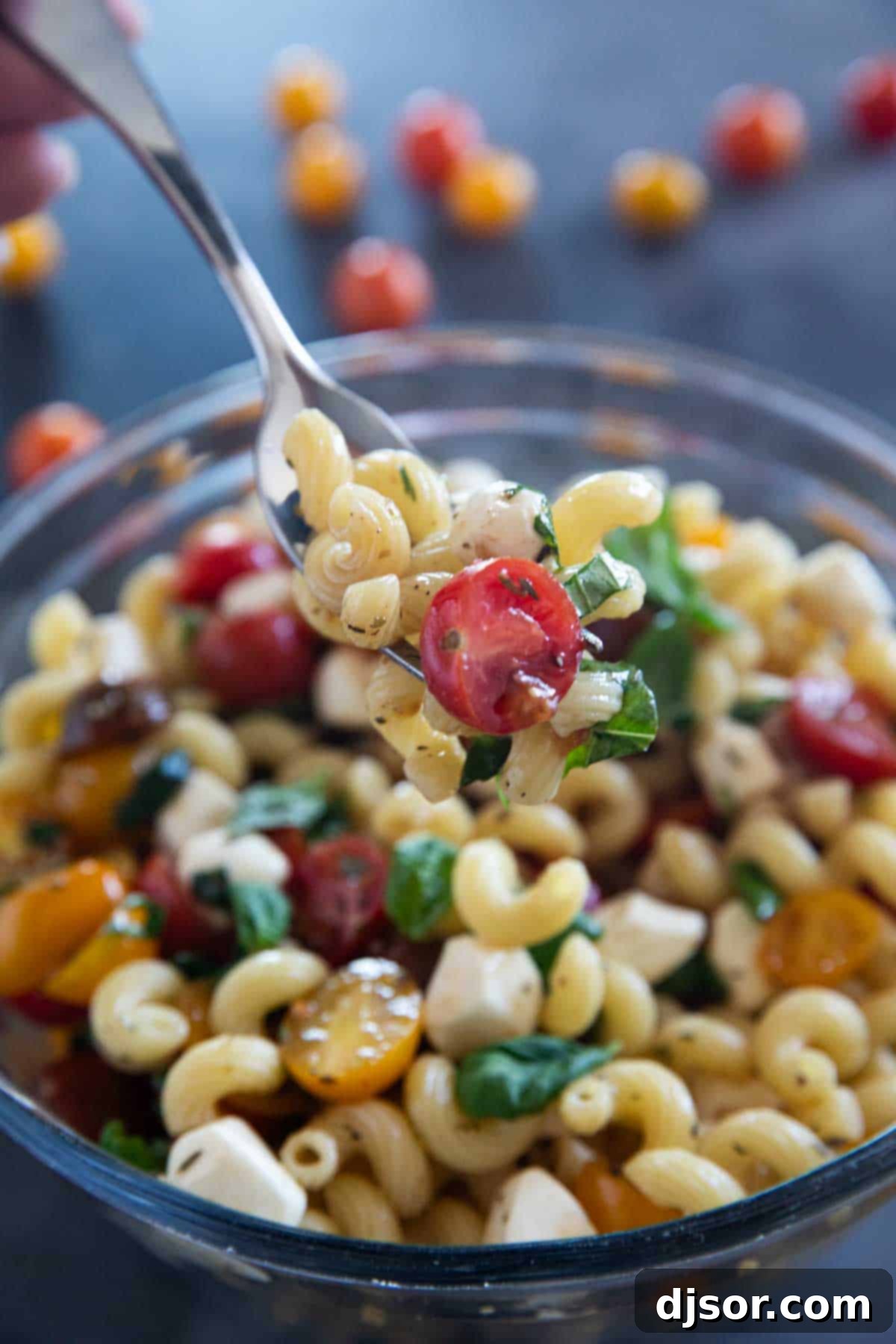 A close-up shot of a fork holding a perfect bite of Caprese Pasta Salad, featuring pasta, a halved cherry tomato, a mozzarella pearl, and a basil leaf.