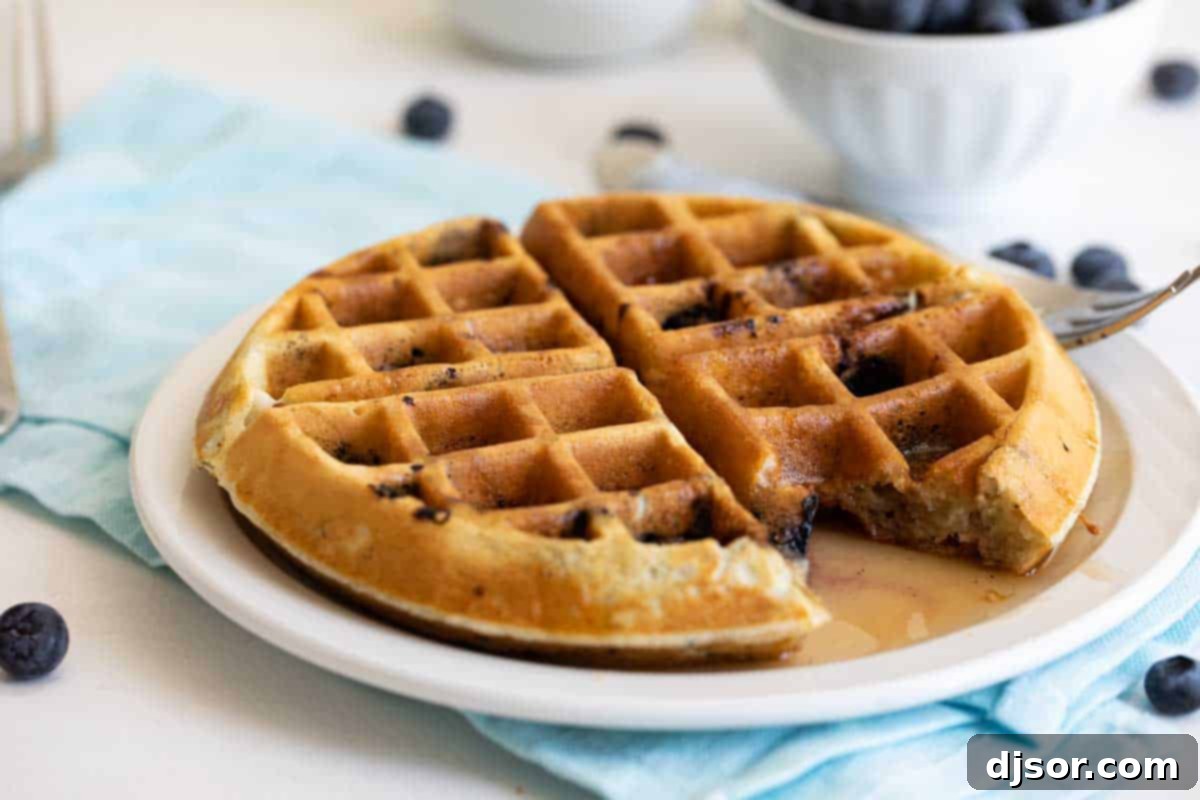 Bursting with blueberry flavor, these tender Blueberry Waffles are a breakfast the whole family loves. They are fast, easy, and the perfect way to make any morning feel special. blueberry waffle on a white plate with a fork and a bite taken from the waffle, showing its fluffy texture
