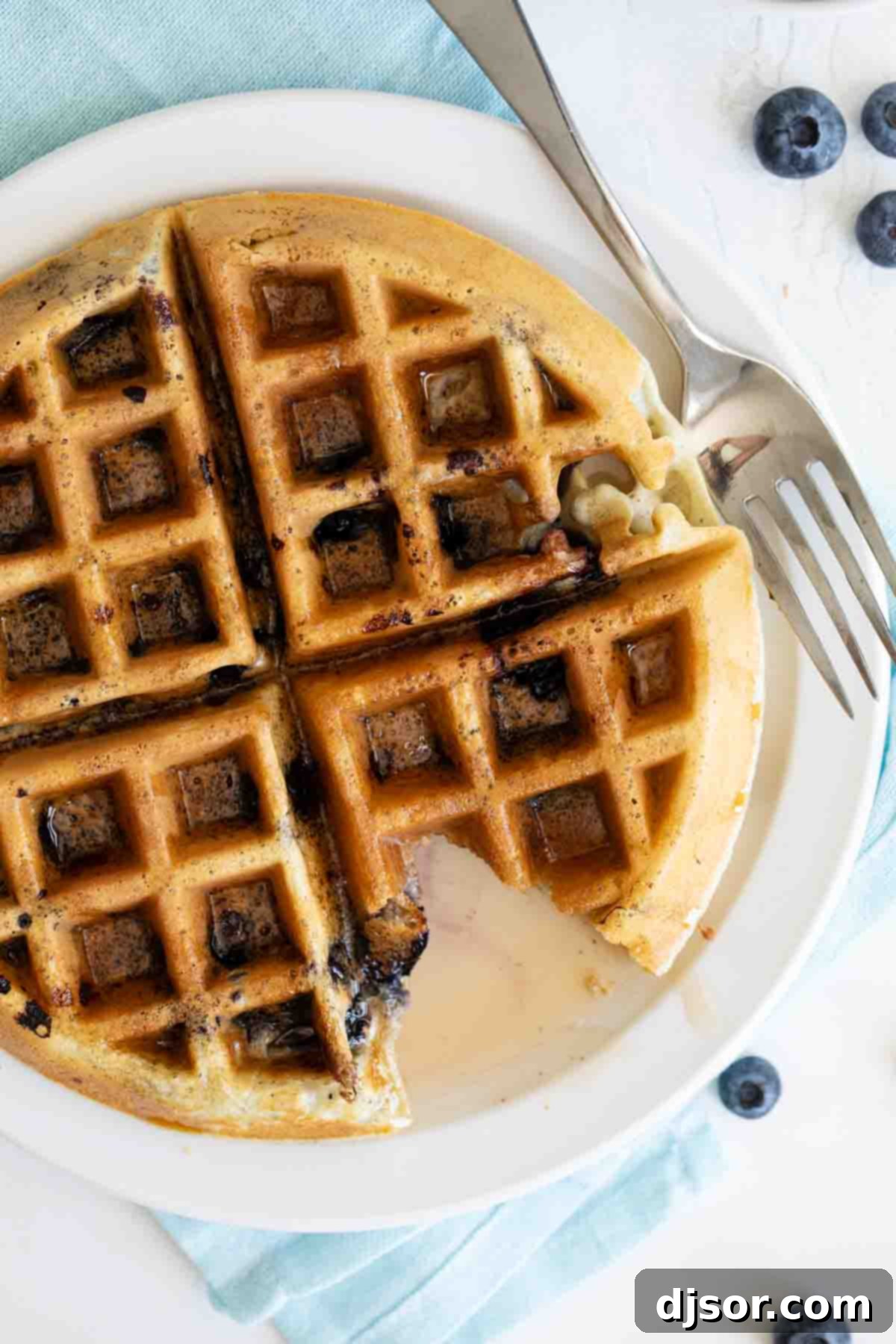 Bursting with blueberry flavor, these tender Blueberry Waffles are a breakfast the whole family loves. They are fast, easy, and the perfect way to make any morning feel special. overhead view of blueberry waffle with syrup with a bite taken, showcasing golden edges