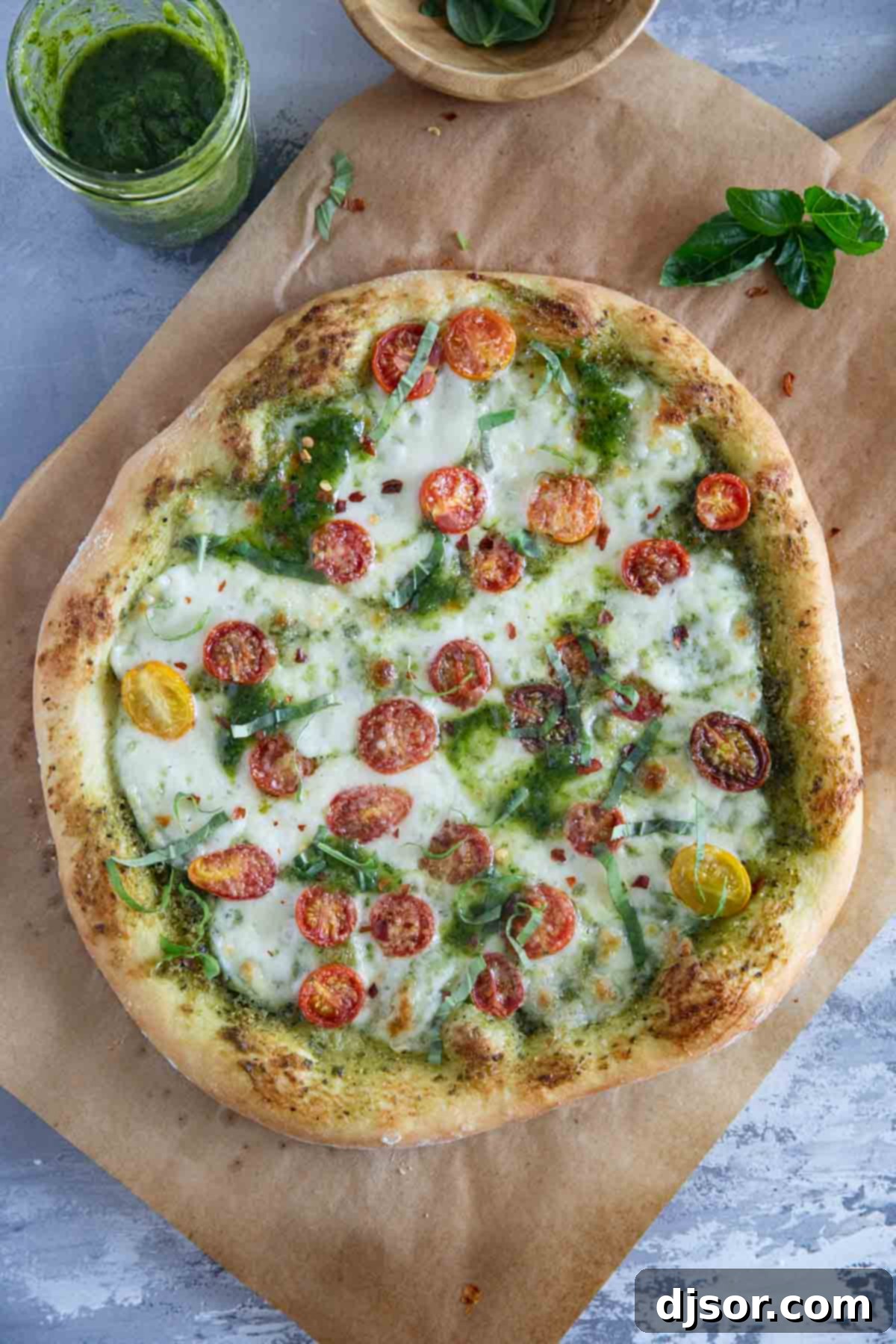 A delicious homemade Pesto Pizza, fresh from the oven, with a perfectly golden-brown crust, melted mozzarella, roasted cherry tomatoes, and a scattering of fresh basil leaves. The full, round pizza promises a delightful culinary experience.