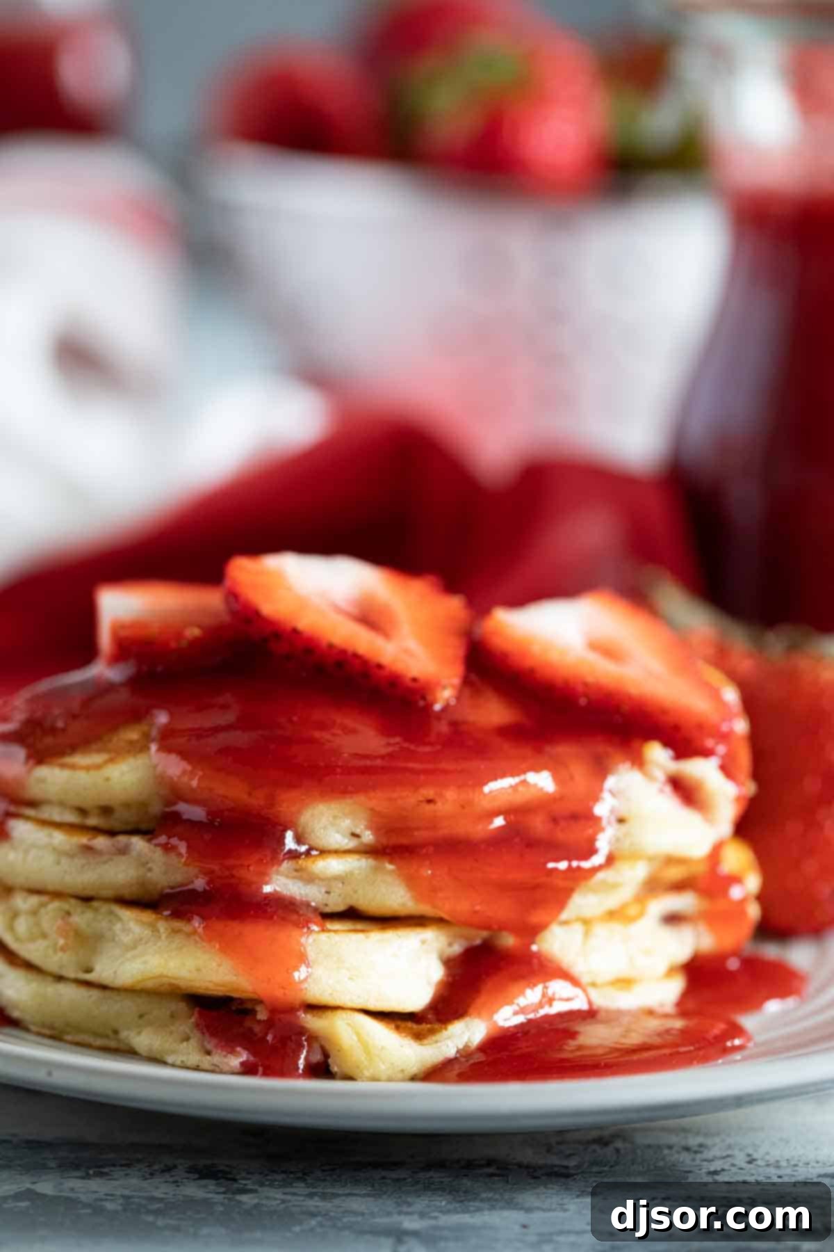 A glorious stack of fluffy strawberry pancakes adorned with fresh strawberry slices and generously drizzled with strawberry syrup, presented on a white plate ready to be enjoyed.