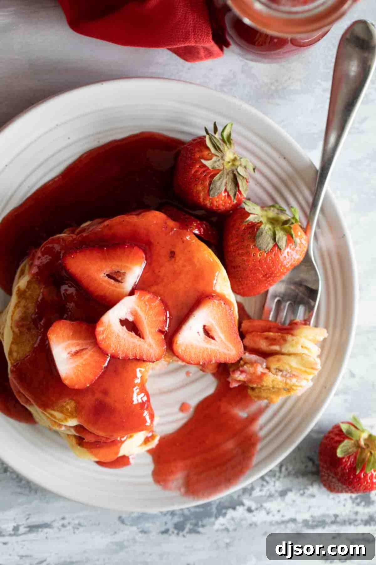 A fork piercing a fluffy strawberry pancake, pulled from a stack, revealing the soft interior and embedded pieces of red strawberry, showcasing its delicious texture.