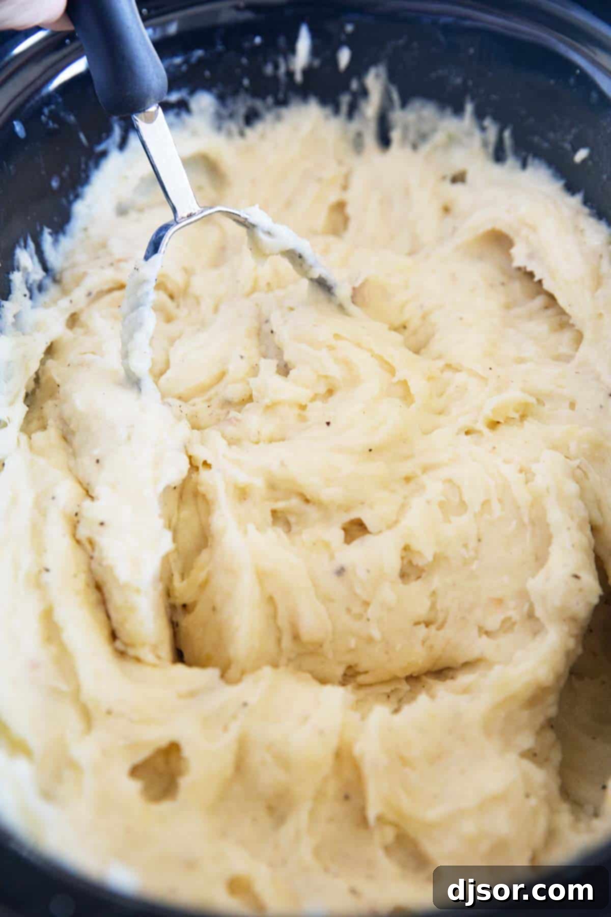 Perfectly Creamy Crockpot Mashed Potatoes 5 Close-up of a potato masher in a slow cooker, actively mashing tender potatoes with added butter and sour cream.