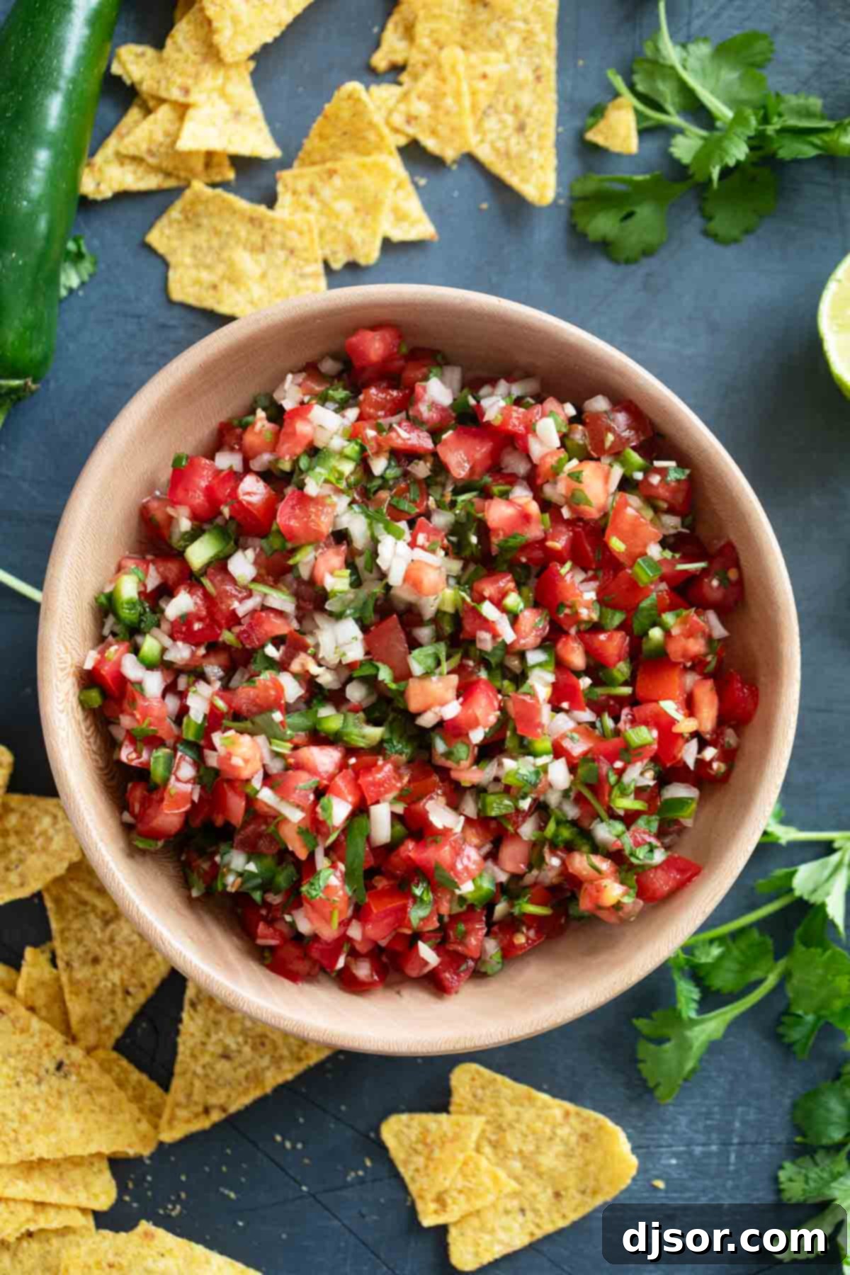 Wooden bowl filled with fresh Pico de Gallo, surrounded by crisp tortilla chips and vibrant, raw vegetables like tomatoes, onions, cilantro, and limes, suggesting the fresh ingredients used.