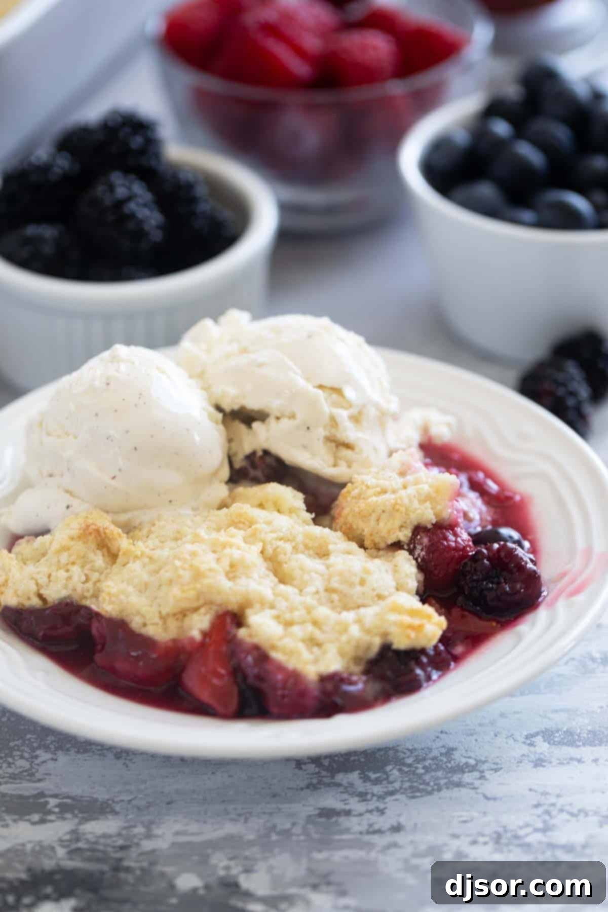 A bowl of freshly baked Berry Cobbler, topped with a scoop of creamy vanilla ice cream, with fresh berries artfully arranged in the background.