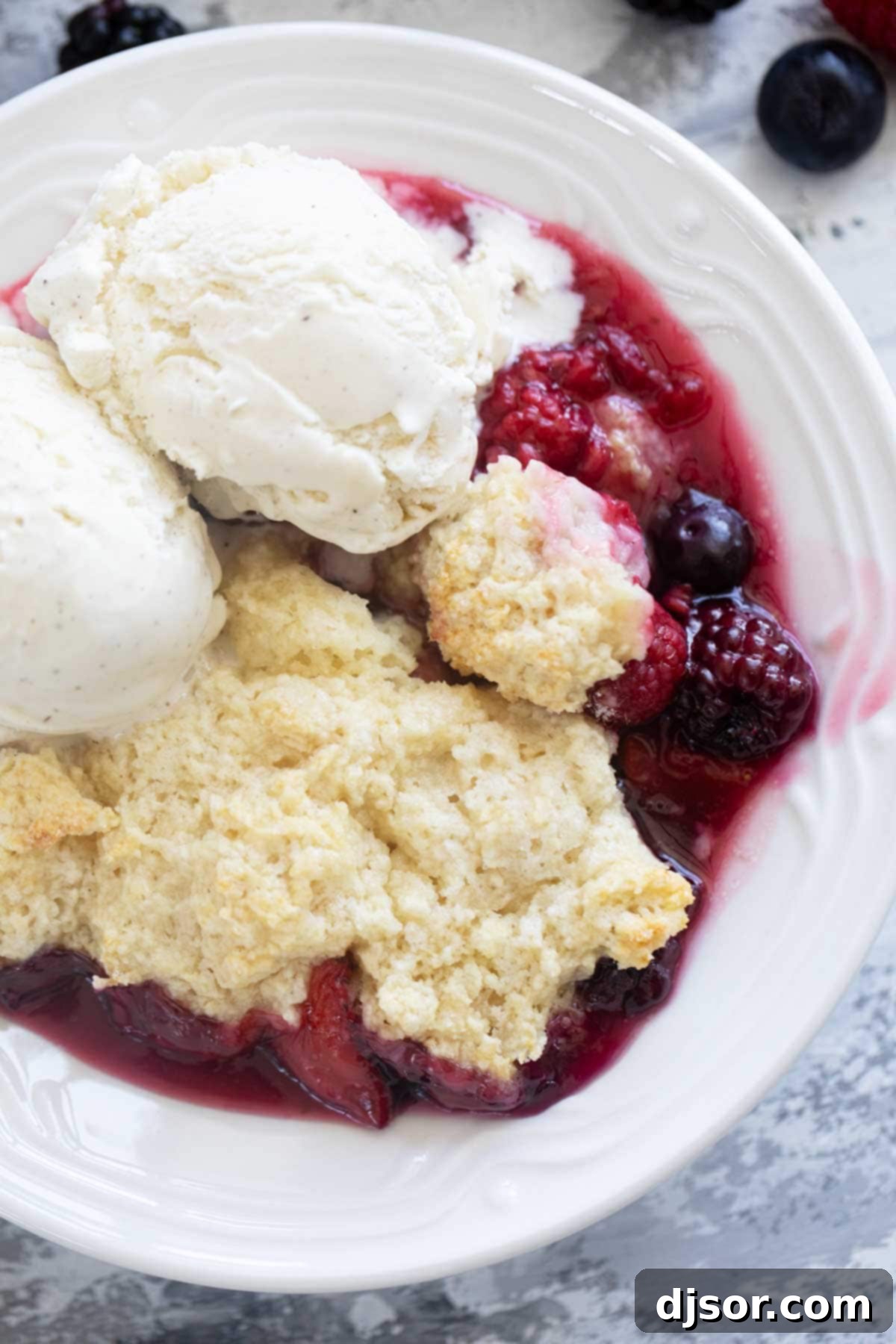 A close-up shot of a bowl of warm Berry Cobbler, generously topped with a scoop of melting vanilla ice cream and garnished with a few fresh berries.