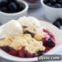 Berry Cobbler in a small bowl, topped with vanilla ice cream with fresh berries in the background.