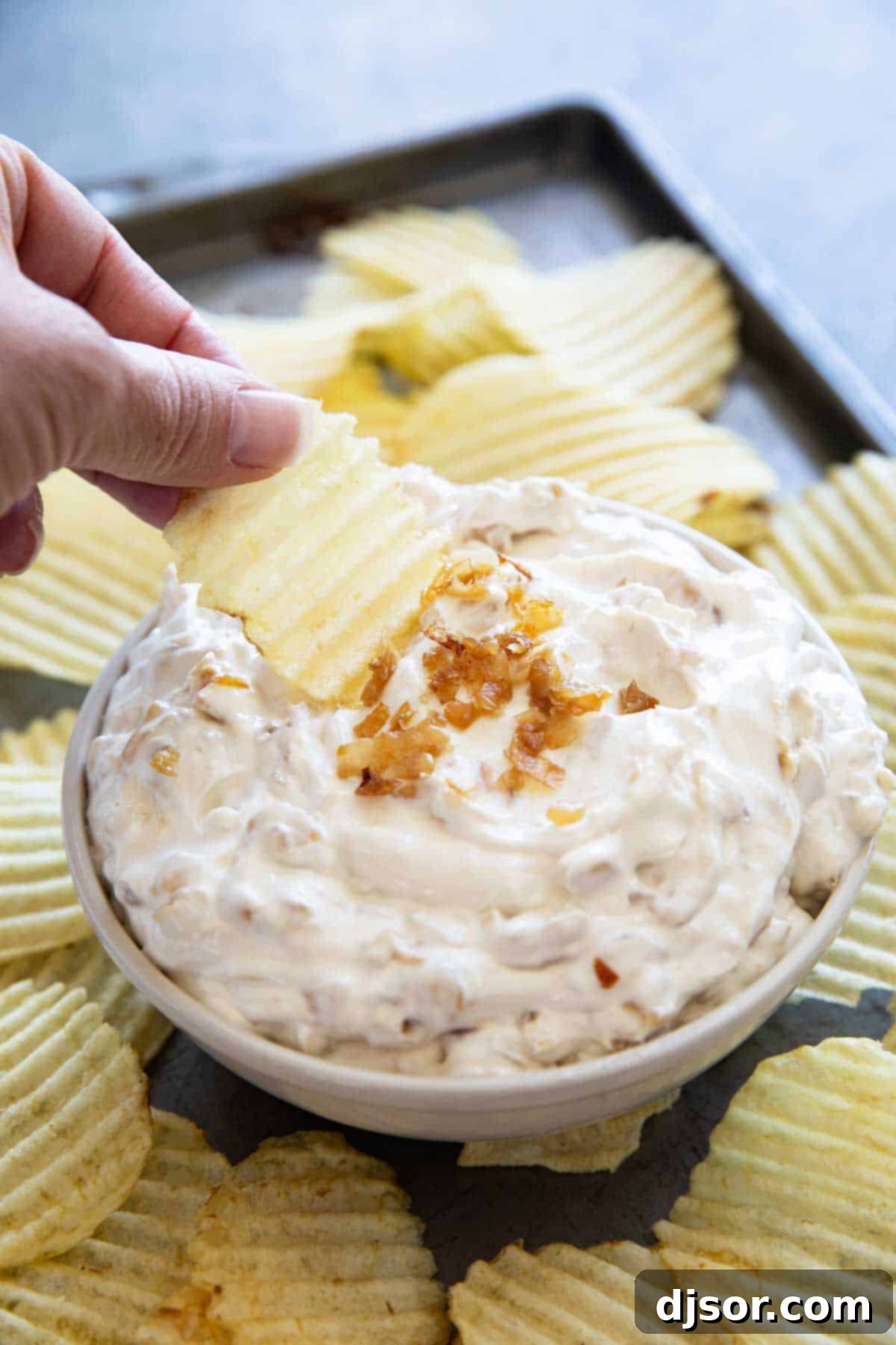 A hand dipping a crispy potato chip into a bowl of creamy French Onion Dip, highlighting its irresistible texture.