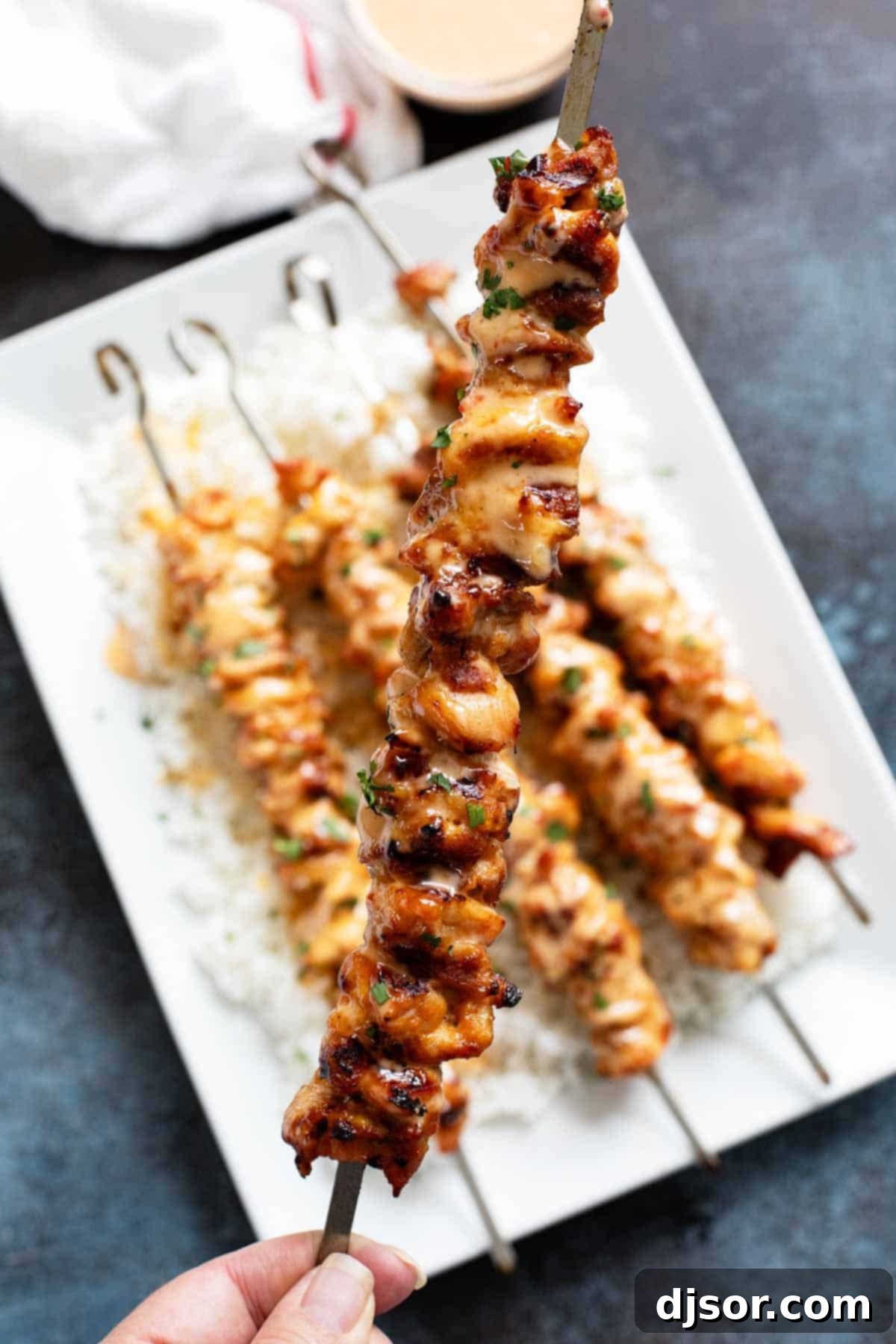 Ultimate Bang Bang Chicken Skewers 7 A hand holding up a single Bang Bang Chicken Skewer, with more skewers laid out on a plate below.