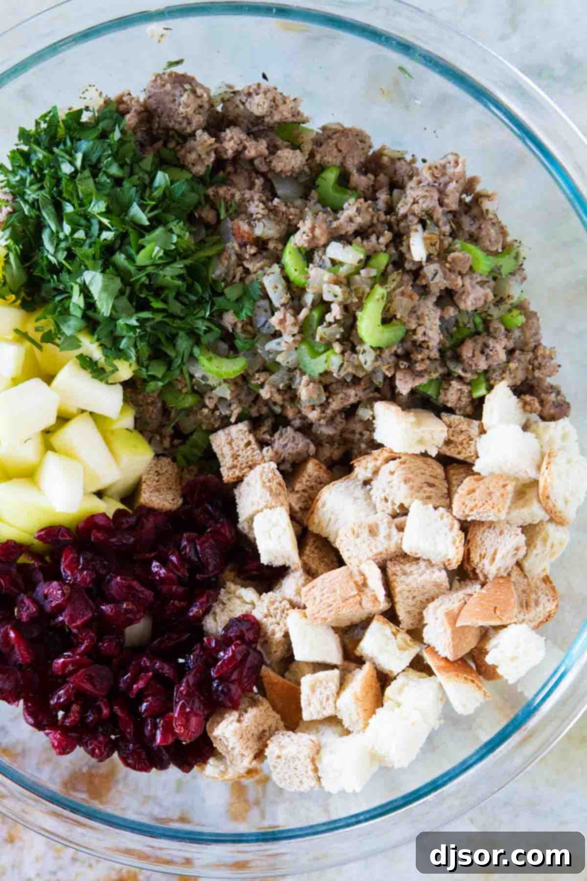 All of the flavors of fall come together in this Sausage Stuffing with Apples and Cranberries for the perfect holiday stuffing recipe. Fresh ingredients laid out for making delicious Sausage Stuffing: bread cubes, turkey sausage, onions, celery, herbs, dried cranberries, and a golden apple.