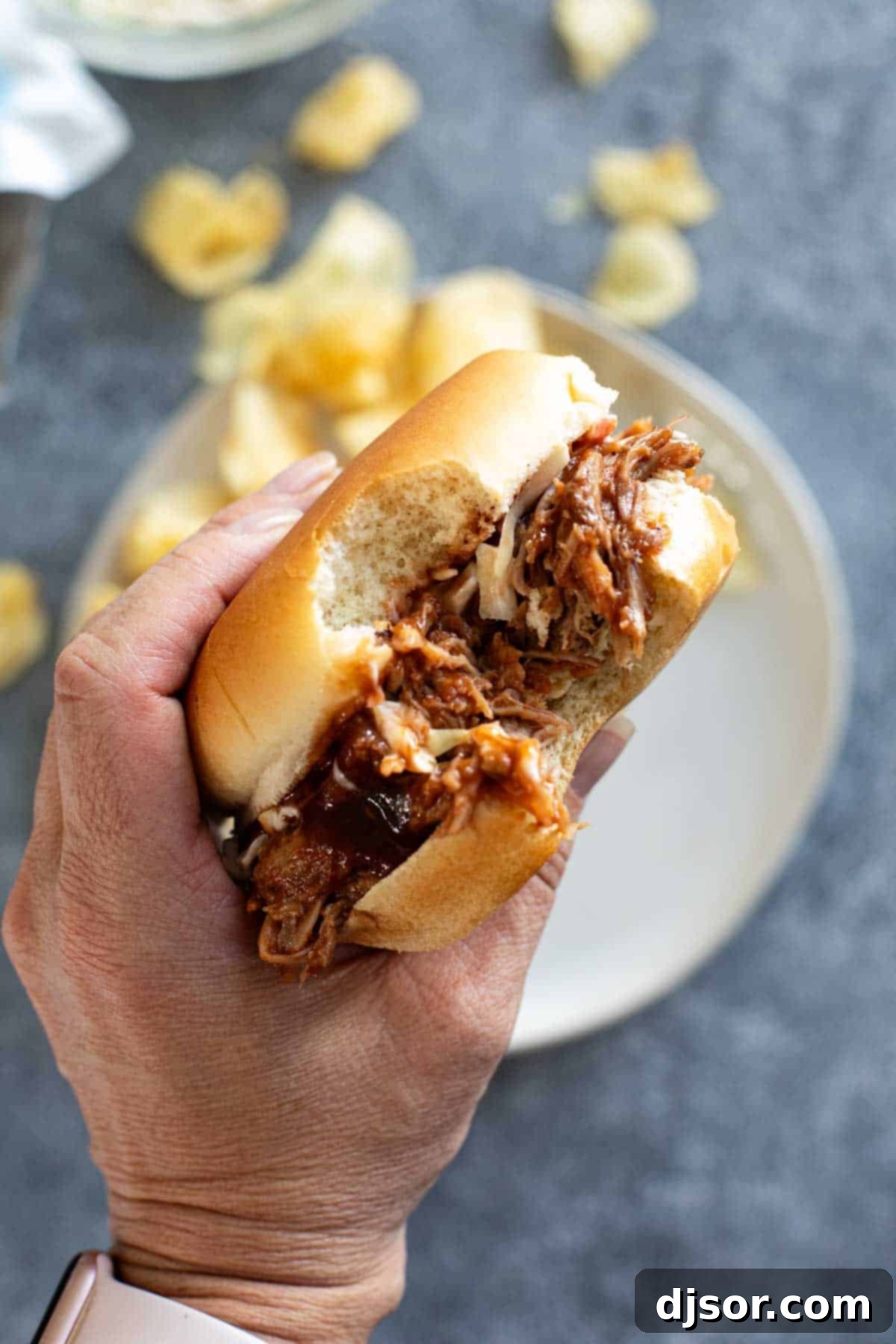 A hand holding a perfectly assembled Slow Cooker Pulled Pork sandwich, complete with a generous filling and a soft bun.