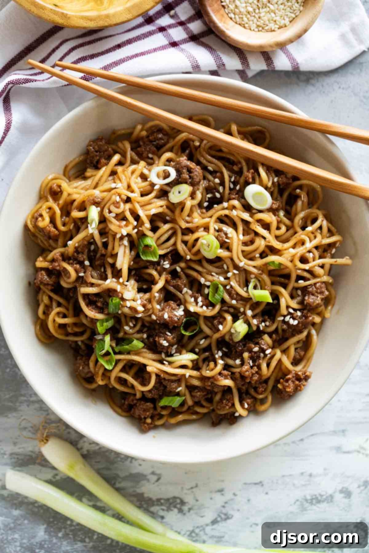 A beautifully presented bowl of Mongolian Ground Beef Noodles, garnished with green onions and sesame seeds, with a pair of chopsticks resting on the side, inviting you to dig in.