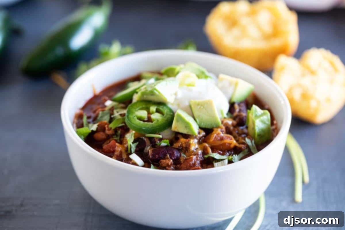 A steaming white bowl of classic crock pot chili, generously topped with shredded cheese, a dollop of sour cream, and fresh diced green onions, sitting on a rustic wooden table.