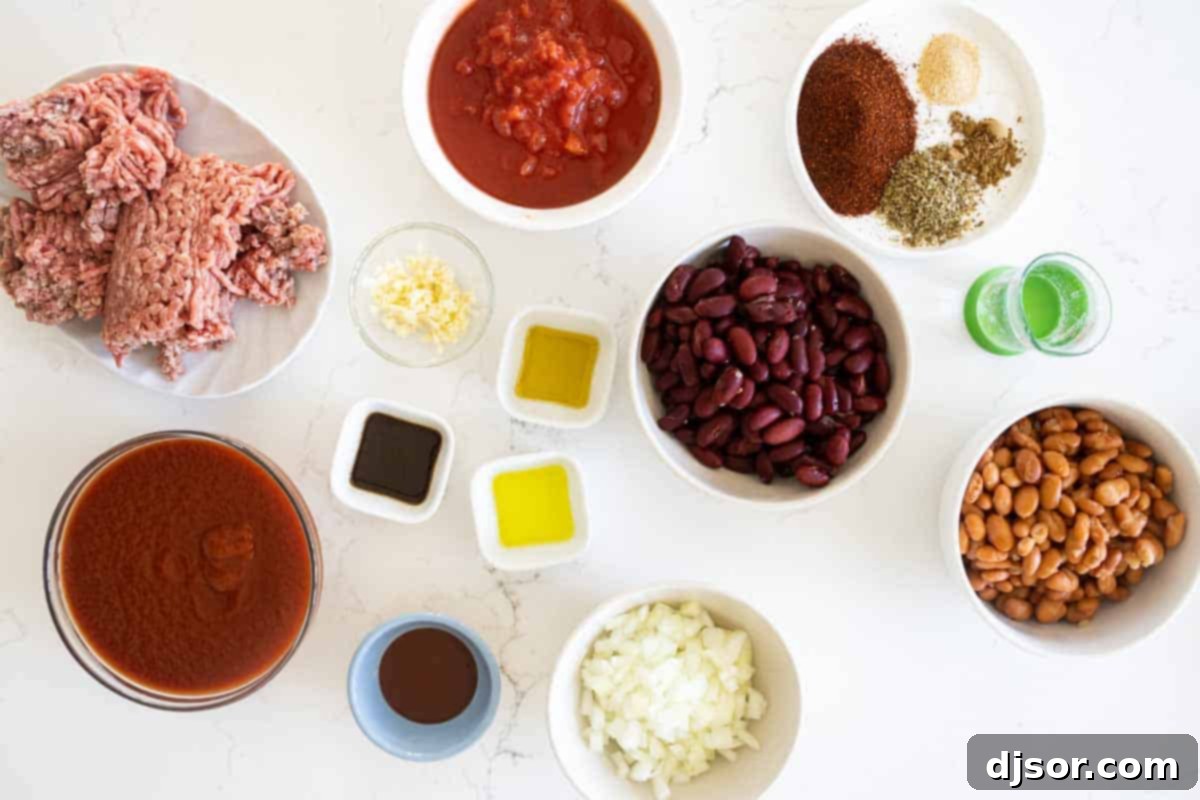 A collection of fresh ingredients laid out on a kitchen counter, including olive oil, onions, garlic, ground beef, various canned beans, diced tomatoes, tomato sauce, and a selection of spices, ready for making crock pot chili.
