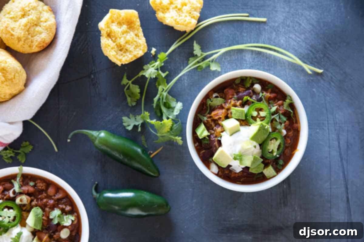 Overhead shot of a bowl of crock pot chili, artfully topped with sour cream, shredded cheese, and diced avocado, with a golden corn muffin and a spoon resting on a white napkin beside it.