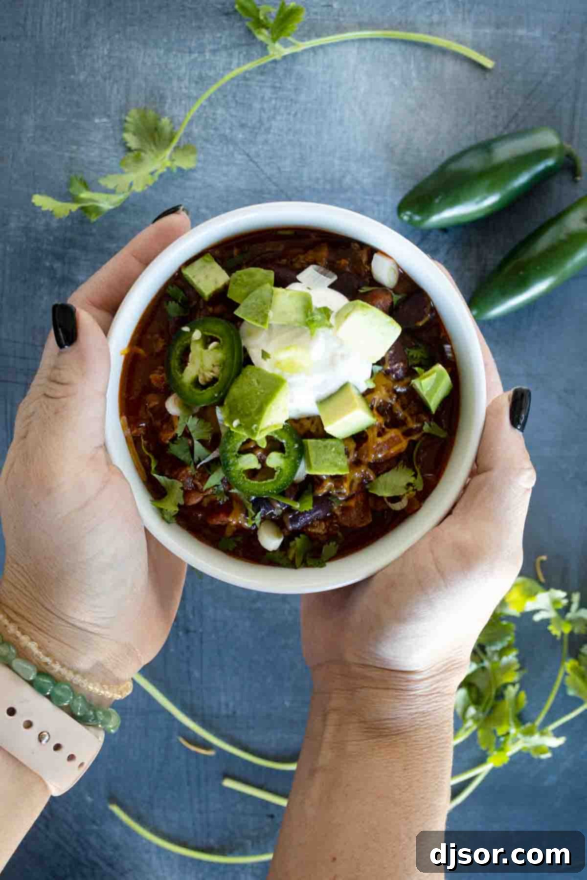 Two hands gently cradling a rustic white bowl filled with steaming, richly colored crock pot chili, emphasizing its comforting appeal.