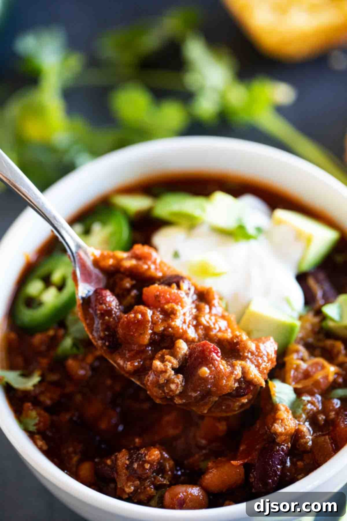 A close-up of a spoonful of crock pot chili, showcasing its rich, thick texture, topped with a dollop of sour cream and fresh diced avocado, ready to be enjoyed.