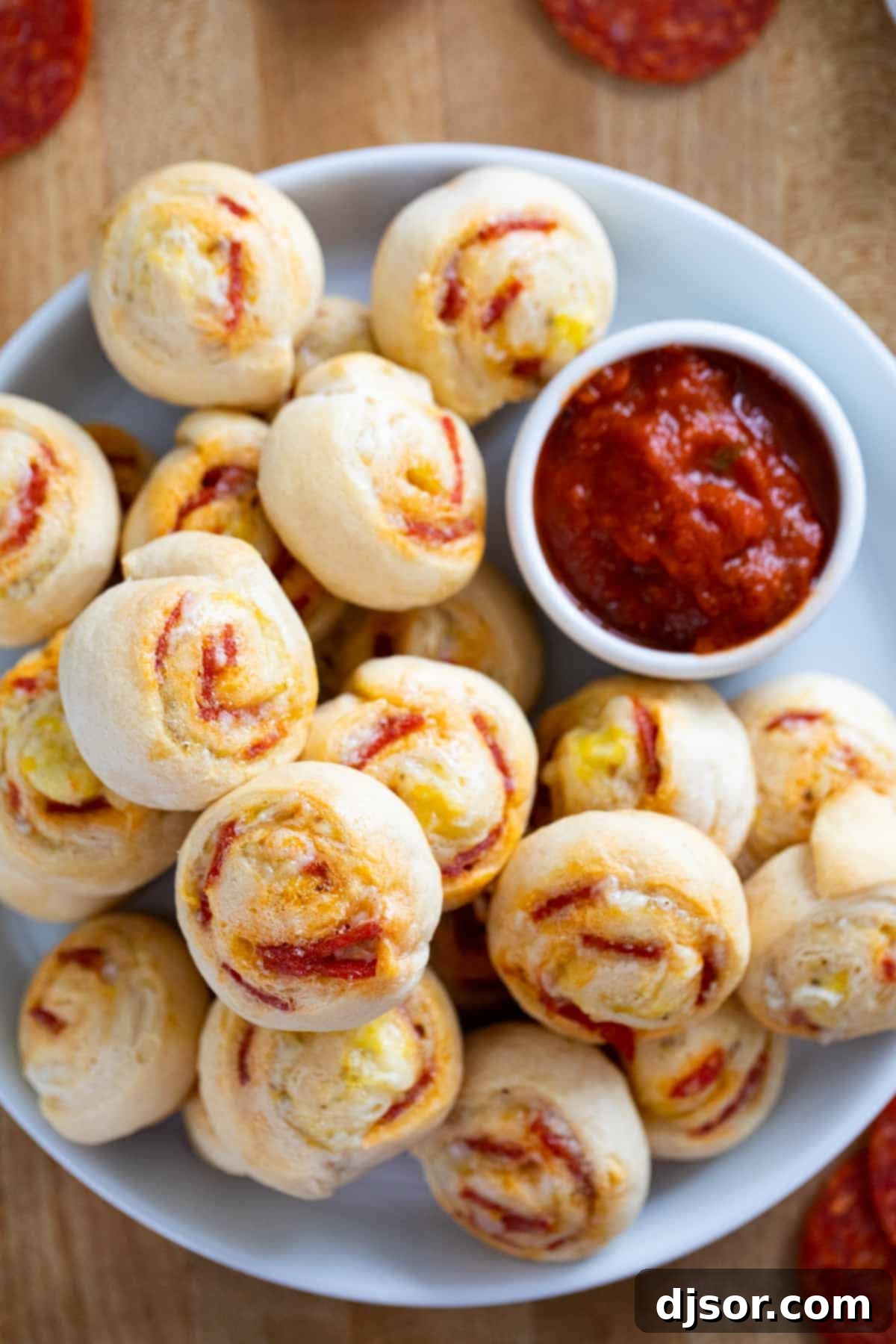 Cheesy Pepperoni Swirls 7 Pepperoni Pinwheels neatly arranged on a plate with a small ceramic bowl of marinara sauce for dipping in the background.