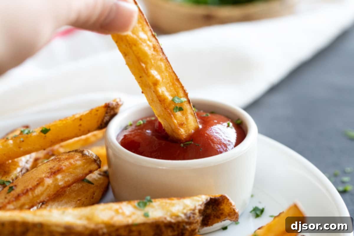 Super crispy and perfectly spiced, these Baked French Fries are the perfect snack or side dish. Forget the deep fryer, these homemade fries are just as good and are better for you! A hand dipping a perfectly golden baked French fry into a small white bowl of red ketchup, ready to eat.