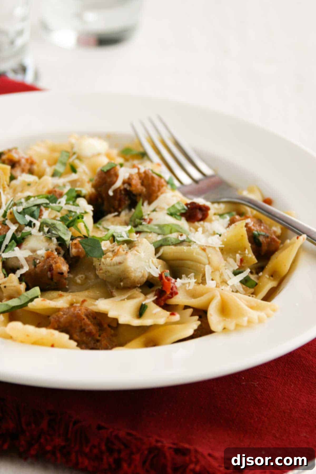 A close-up of a generous serving of pasta with Italian sausage, quartered artichoke hearts, and sliced sun-dried tomatoes in a white bowl. The dish is beautifully garnished with fresh basil leaves and a sprinkle of grated Parmesan cheese, highlighting its vibrant colors and rich textures.