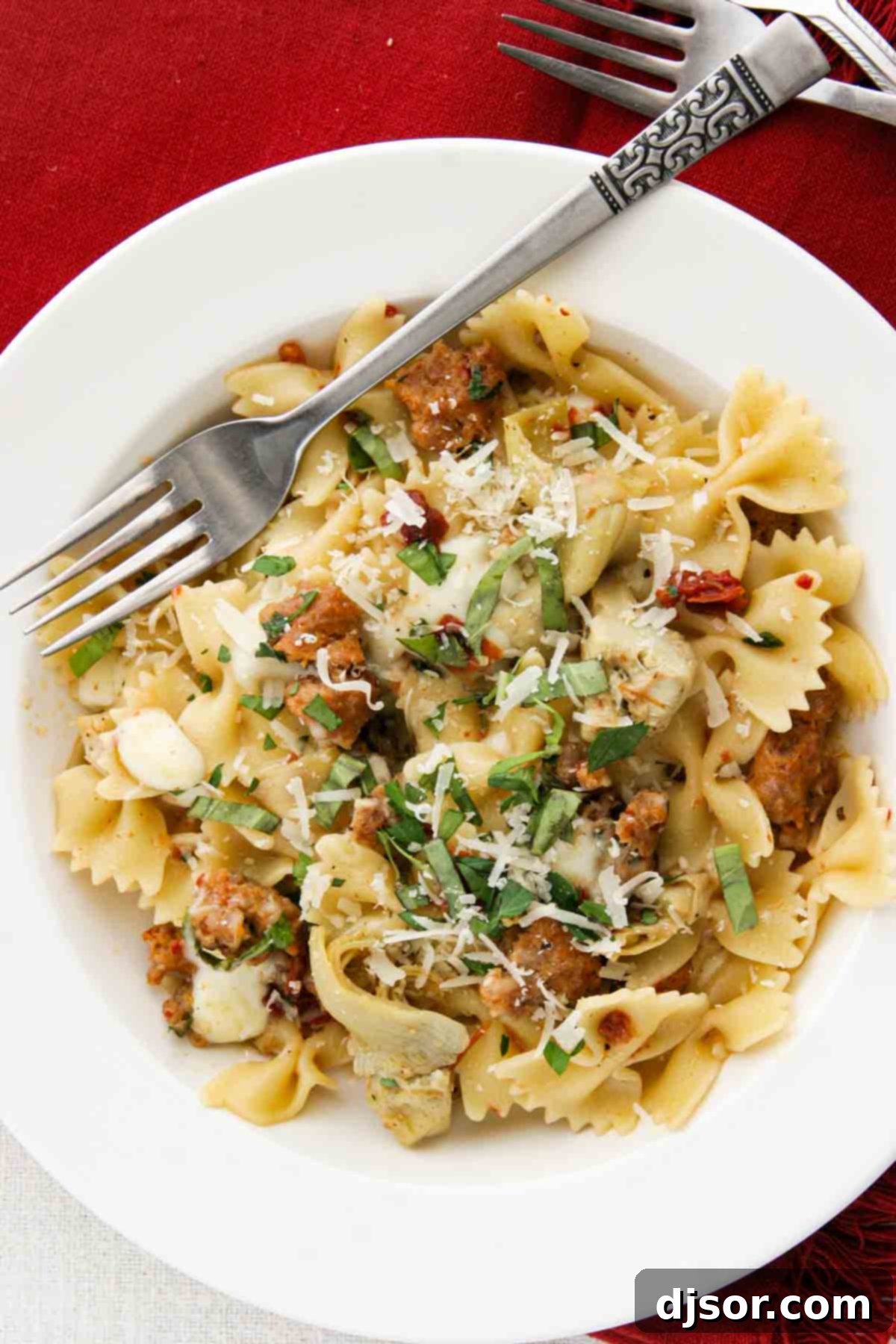 A top-down view of pasta with Italian sausage, artichokes, and sun-dried tomatoes served in a shallow white bowl. The pasta is tossed with the savory components, showcasing the golden-brown sausage, green artichoke pieces, and reddish-brown tomatoes, indicating a well-mixed and flavorful dish.